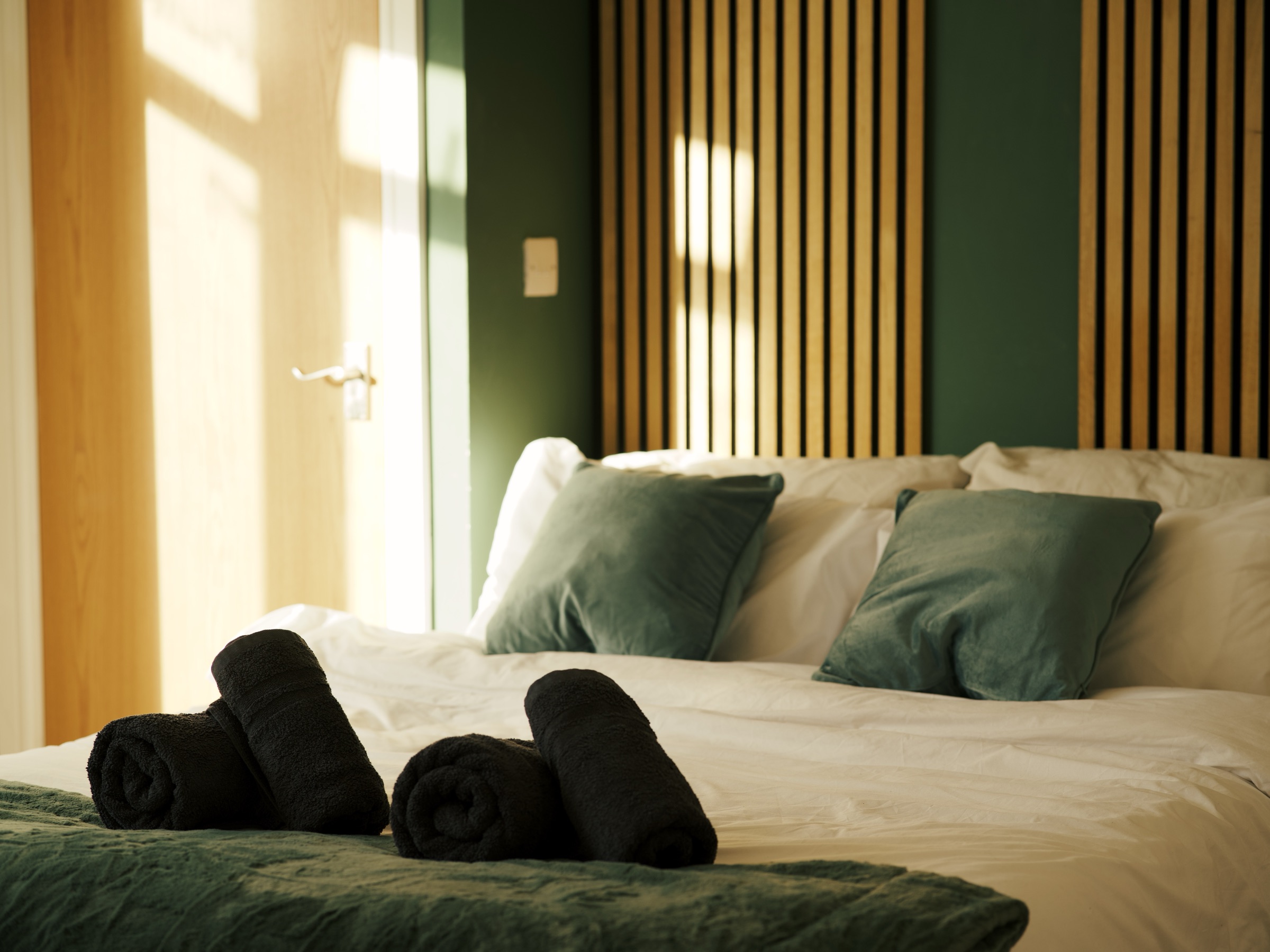 A dressed double bed with rolled towels at its foot, against an oak-slatted headboard wall, in low afternoon light