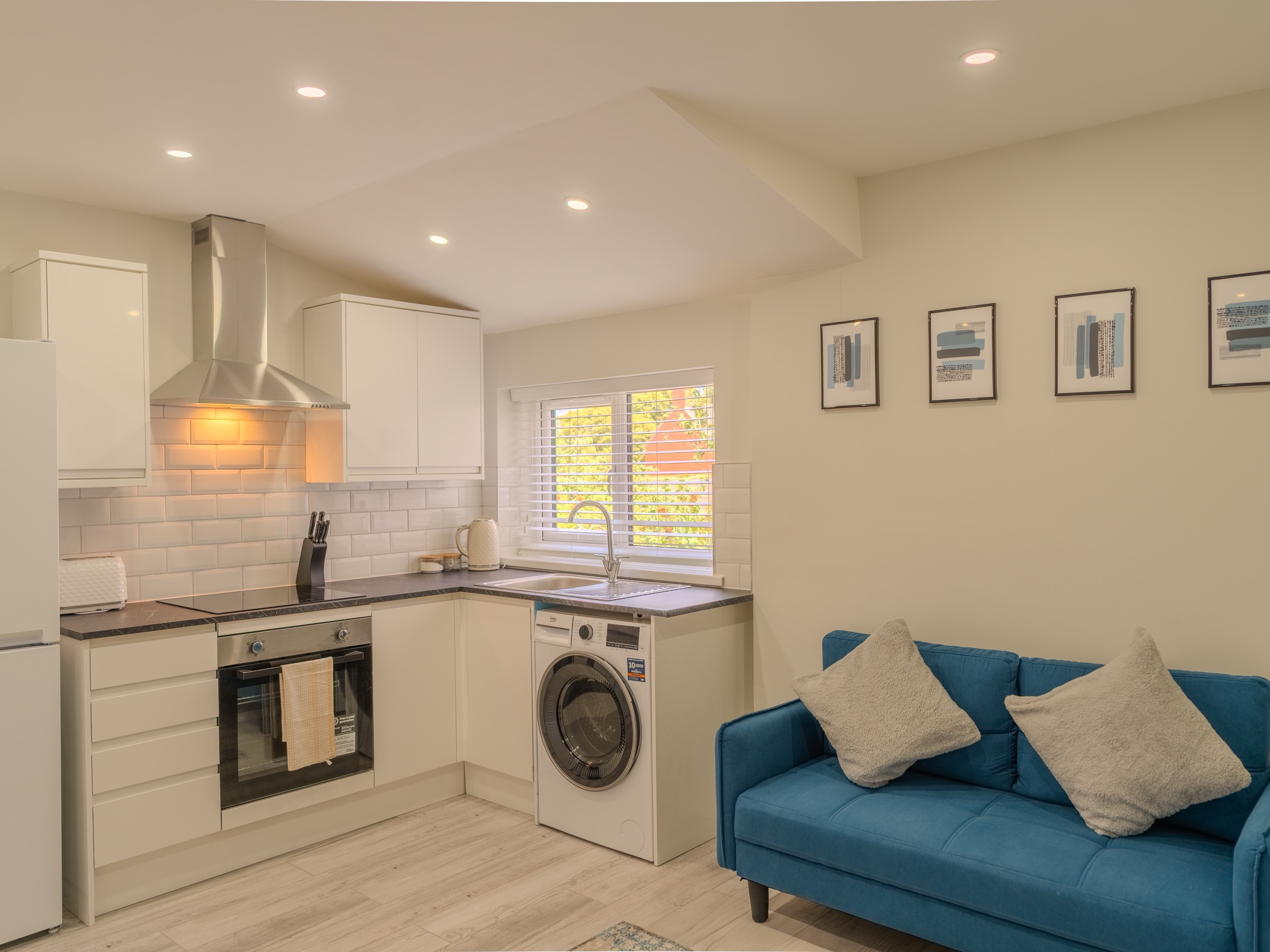 An open-plan kitchen and living room — cream units, a blue two-seater sofa, a row of framed prints