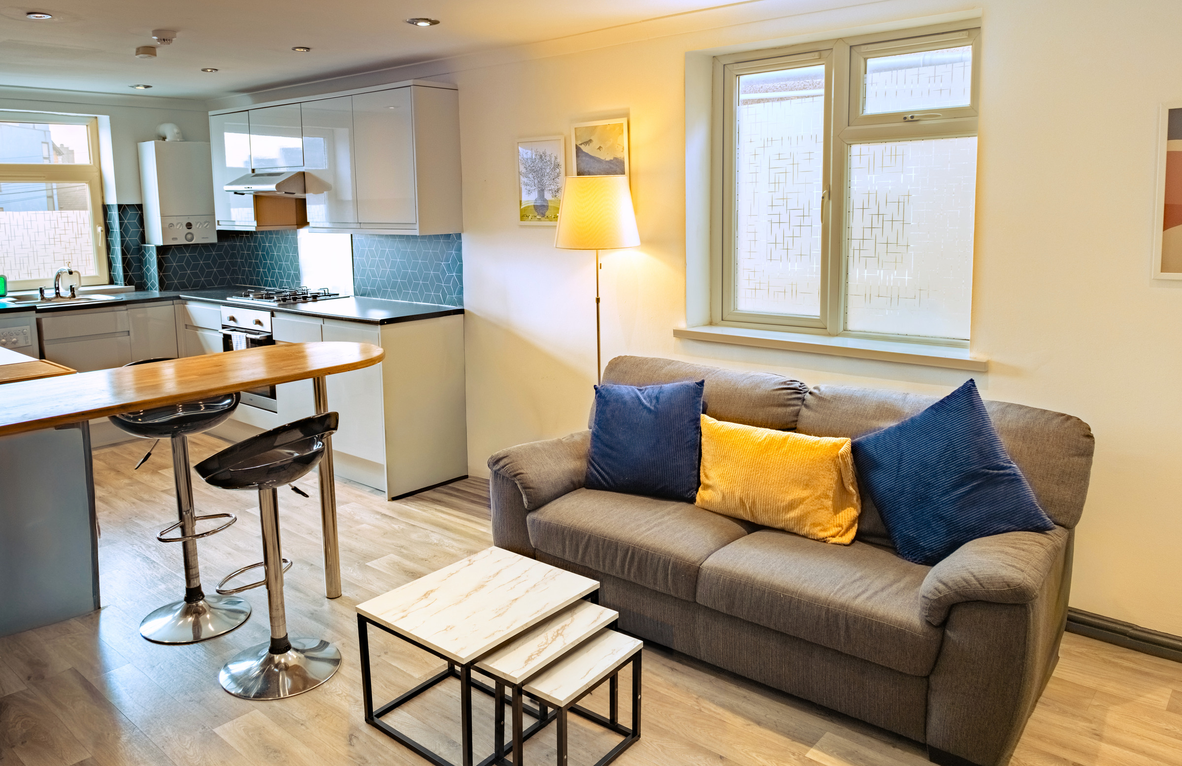 Grey sofa with blue and yellow cushions beside a breakfast counter and fitted kitchen.
