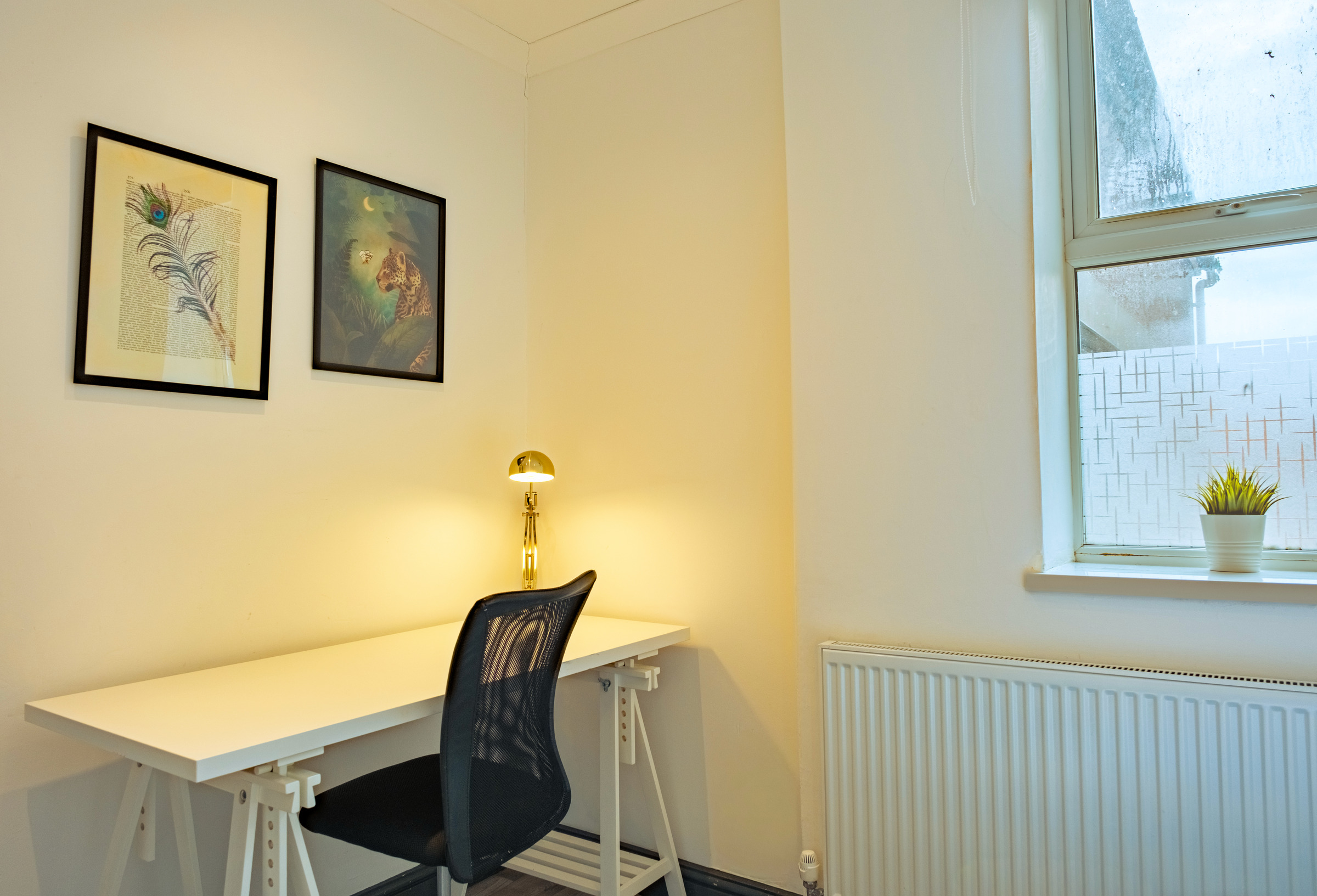 Desk and dark chair beneath two framed pictures beside a daylit bedroom window.