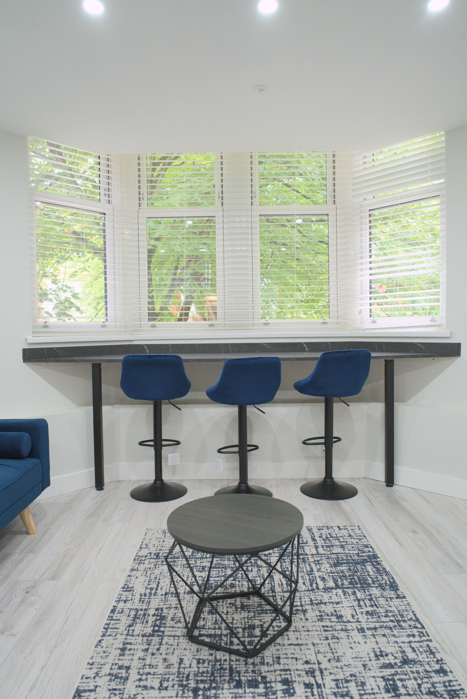 Three blue stools beside a large bay window overlooking trees, with a rug and coffee table.