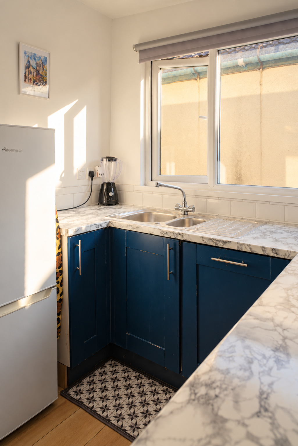 A compact blue kitchen with marble-pattern worktops, double sink and warm sunlight across the fridge.