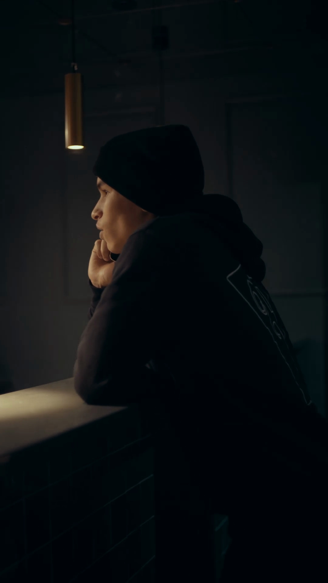 A quiet profile in a black hooded garment beside a warm hanging light, from our finished brand film.