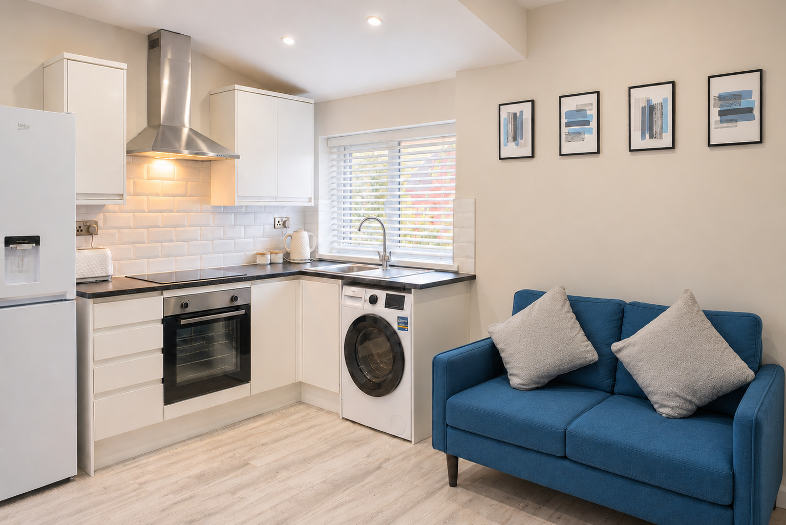 A blue two-seat sofa beside a white kitchen with oven, fridge and washing machine in a compact studio.