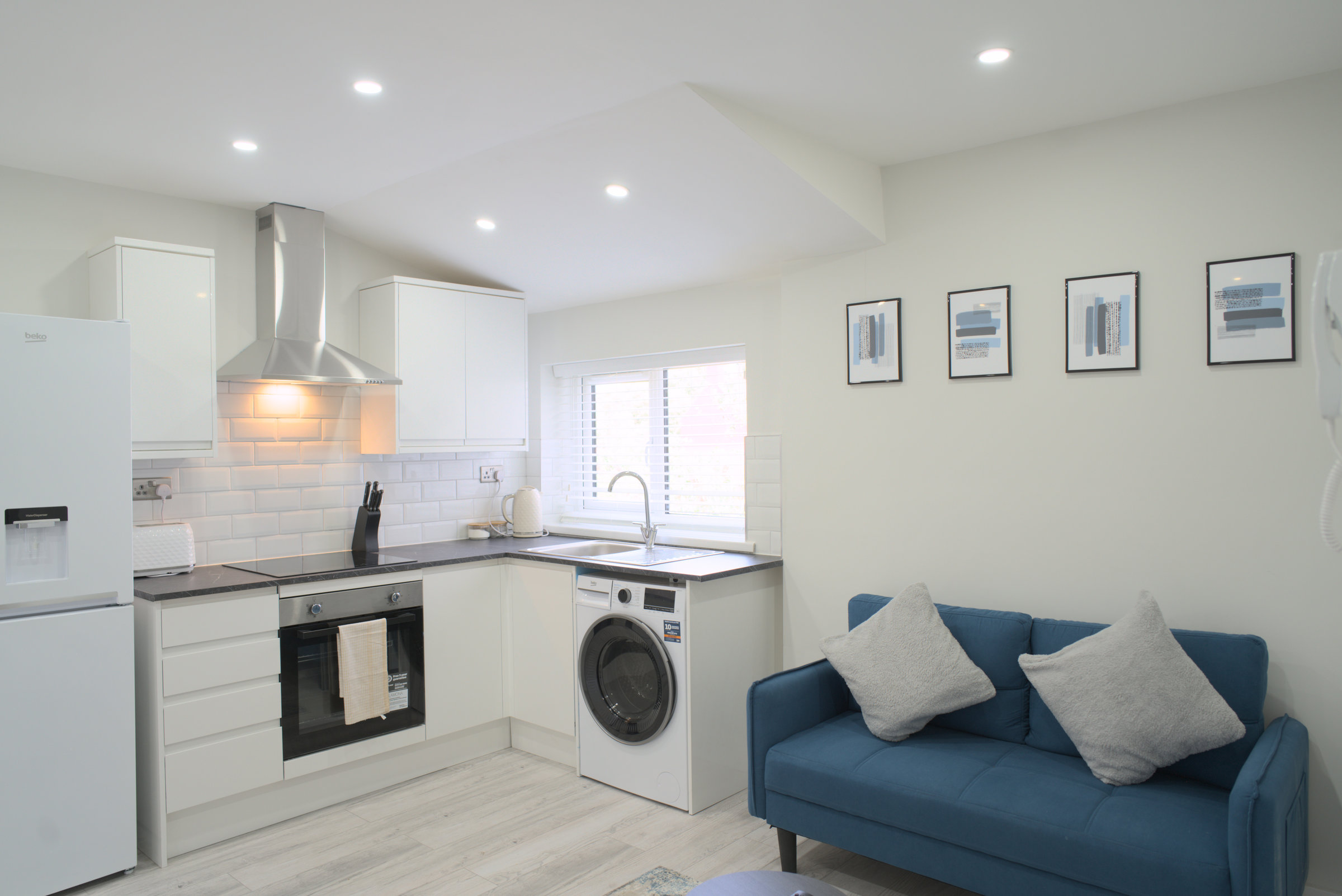 Open-plan apartment with blue sofa, white kitchen and daylight through a street-facing window.