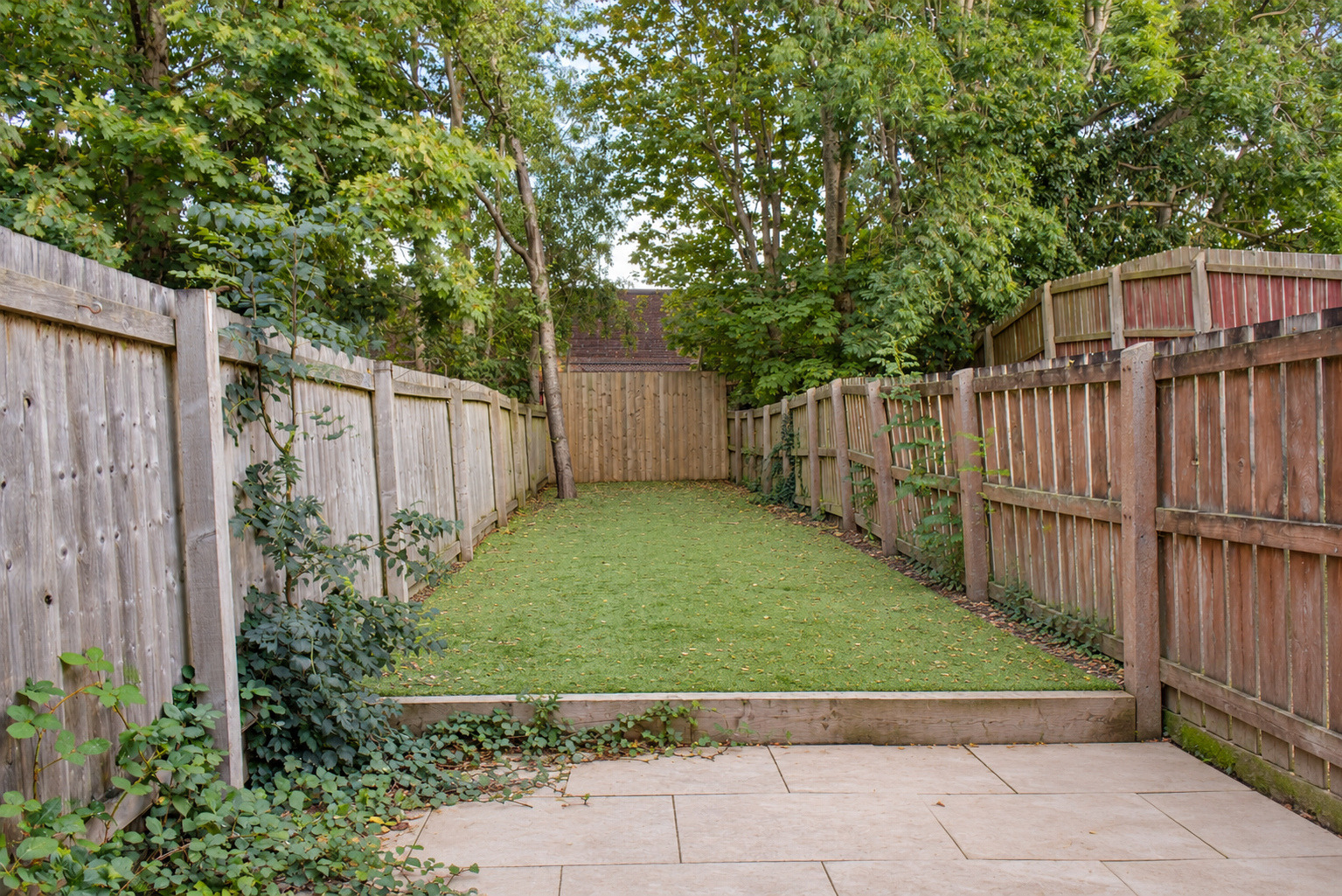 Paved terrace leading to a narrow lawn with timber fencing and established trees.