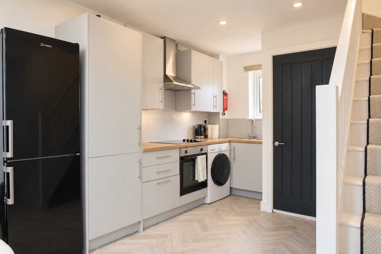 White kitchen cabinets, a black fridge, oven and washing machine beside the stairs.