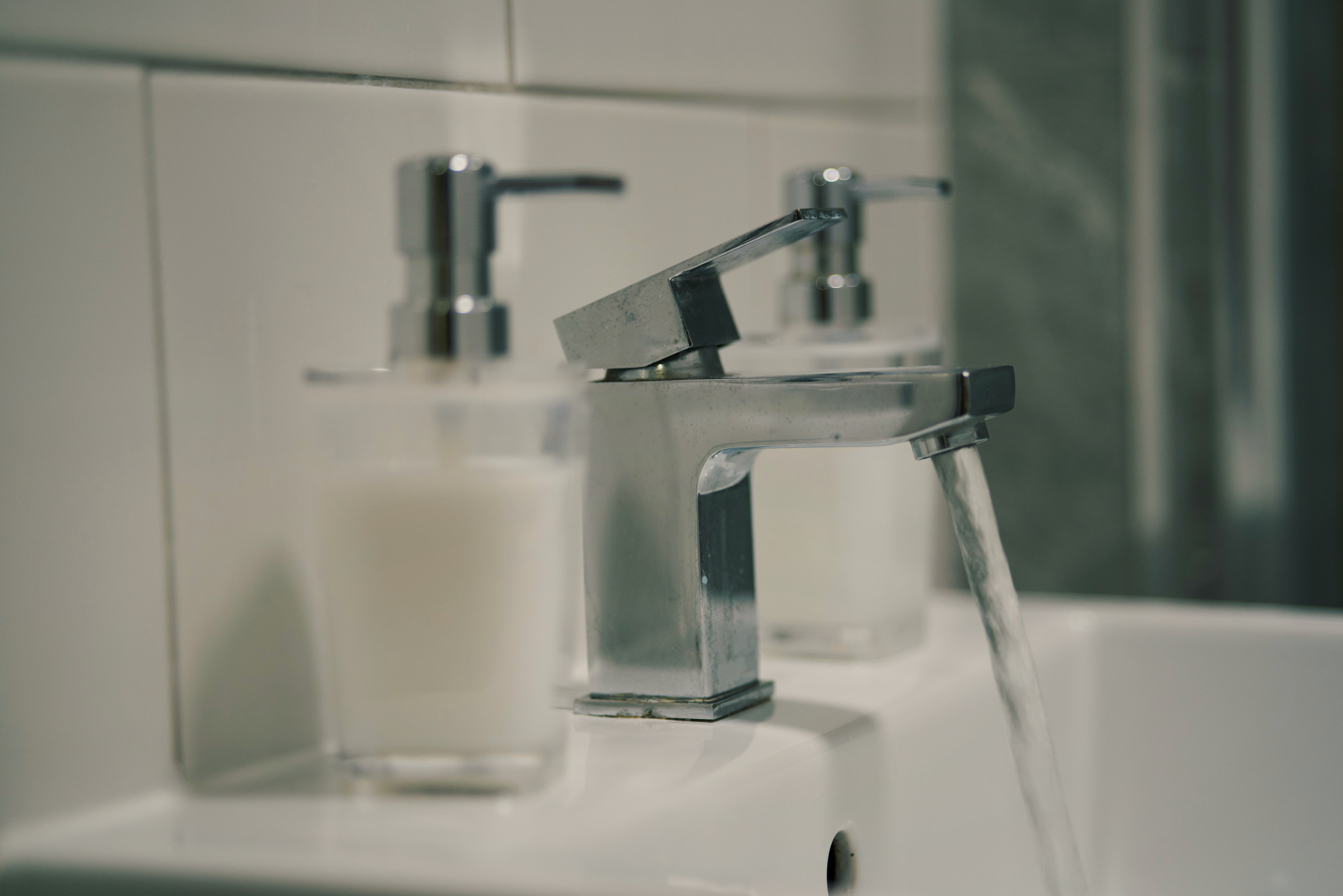 Chrome tap, clear soap dispenser and white basin in a bathroom detail.