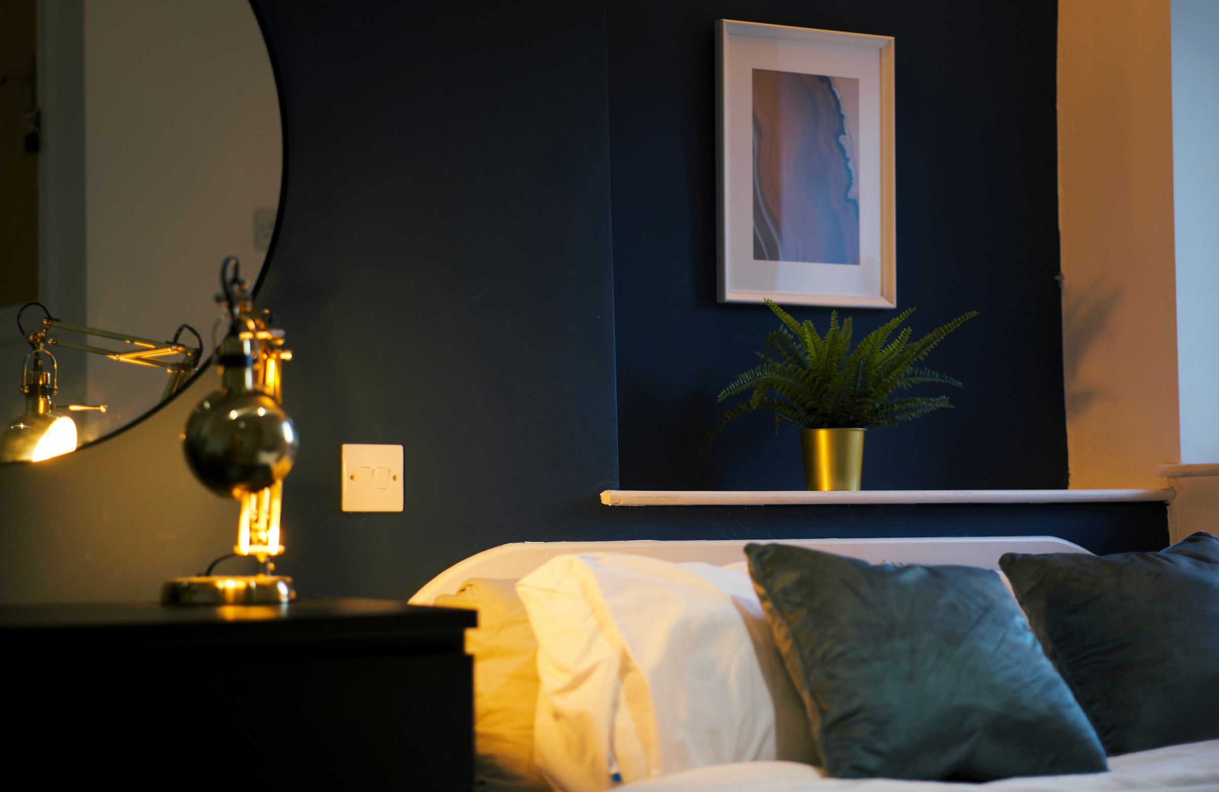 Warm brass bedside light, white pillows and a small plant against the navy bedroom wall.