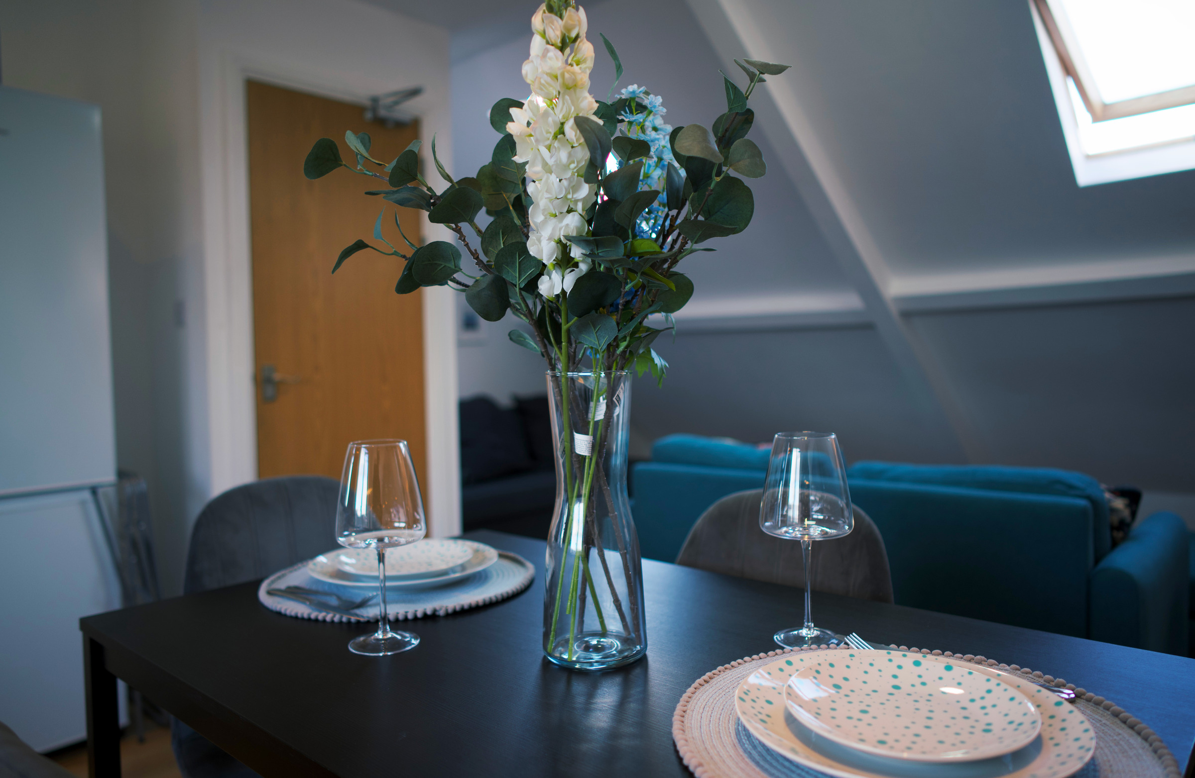 Dark dining table with place settings and flowers beneath a skylight.