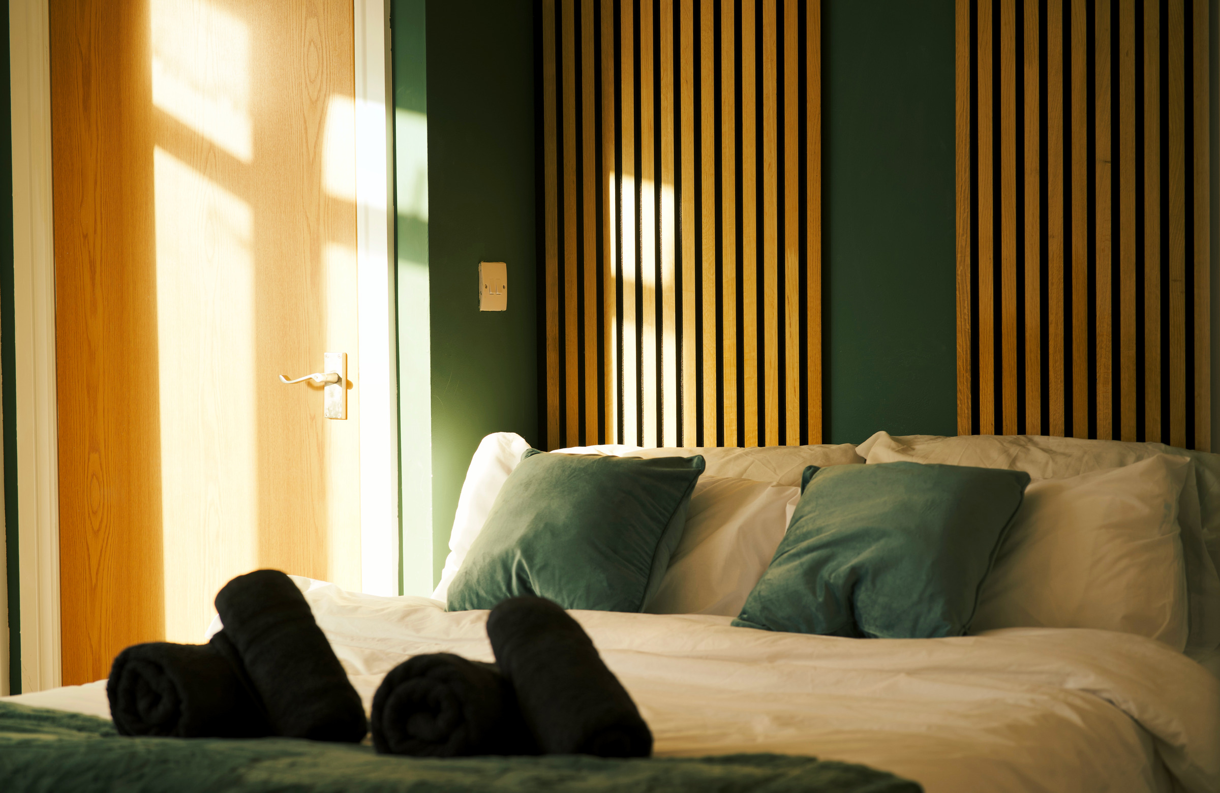Directional sunlight across green bedding, rolled towels and the slatted headboard wall.