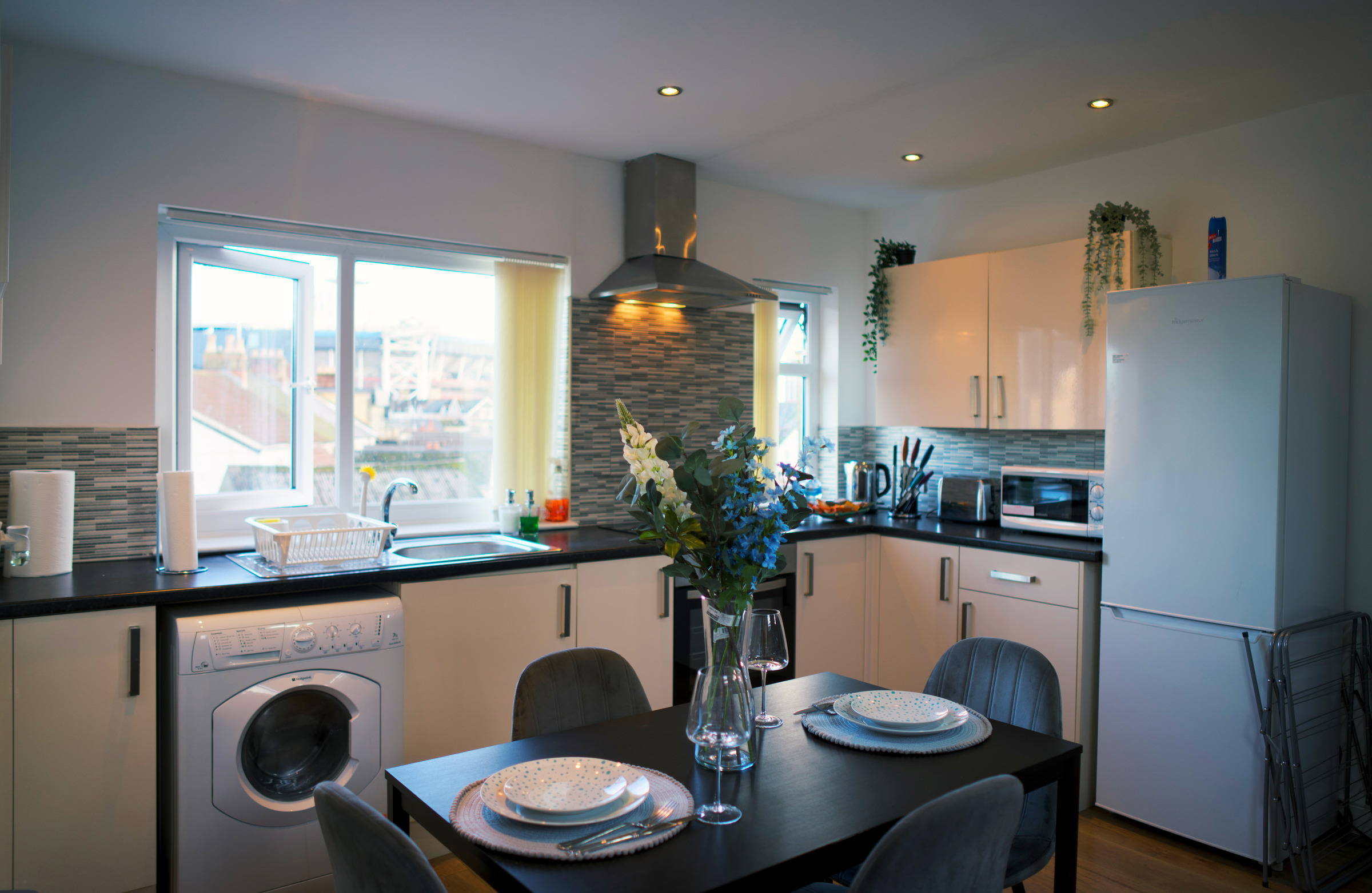 A set dining table beside a pale-blue fitted kitchen and large apartment window.
