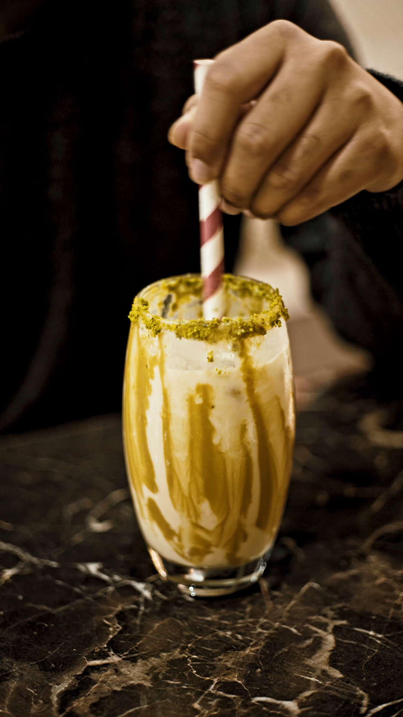 A hand stirring a green-and-cream drink in a glass on a marble restaurant table.
