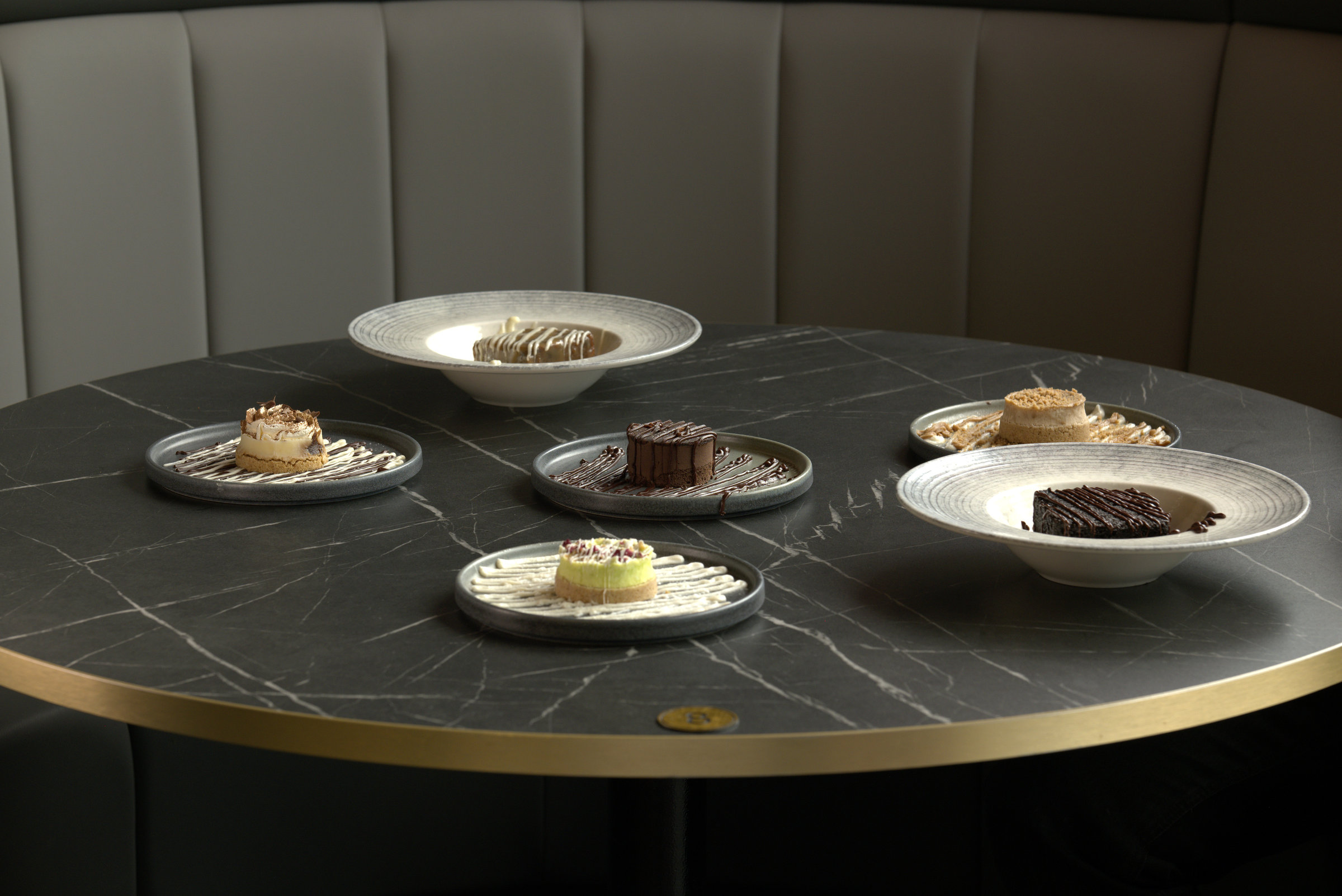 Six desserts arranged across a round marble table in a restaurant booth.
