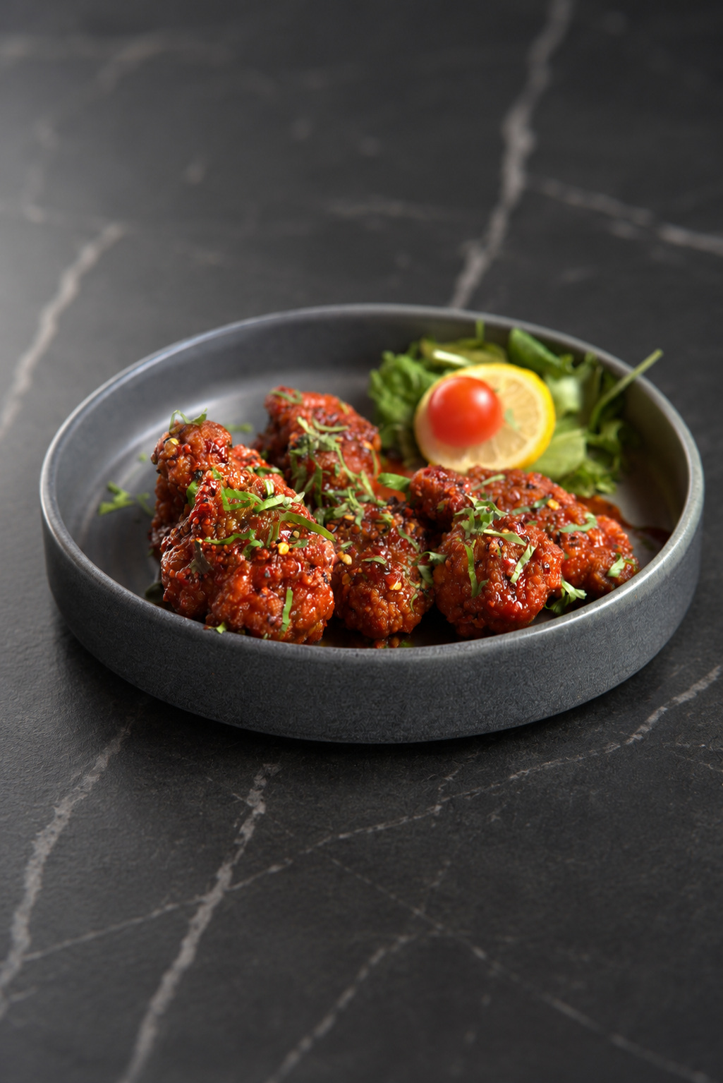 A shallow charcoal bowl of glazed chicken with herbs, salad, lemon and tomato on a dark marble table.