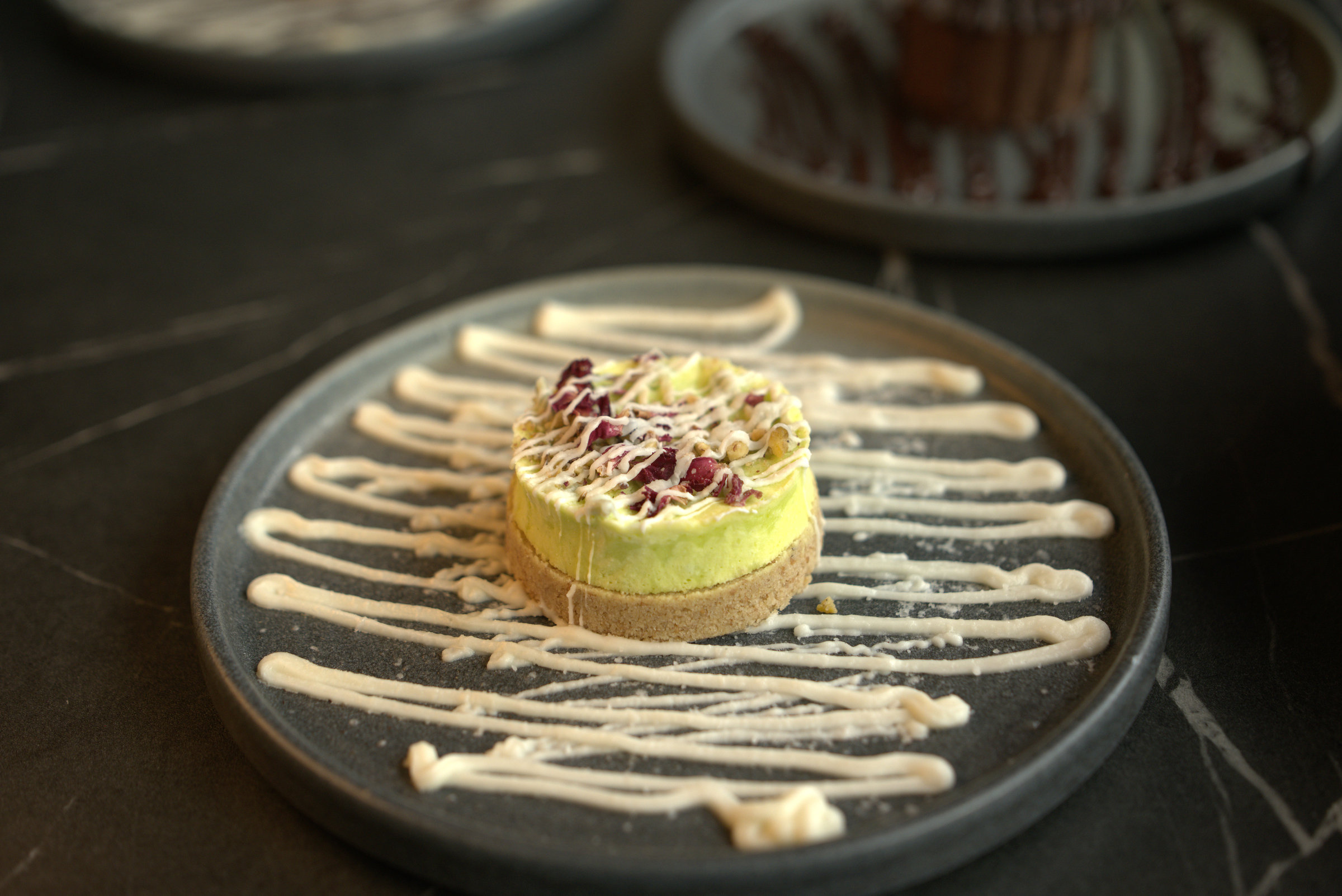 A green layered dessert with cream drizzle and dried petals on a dark ceramic plate.