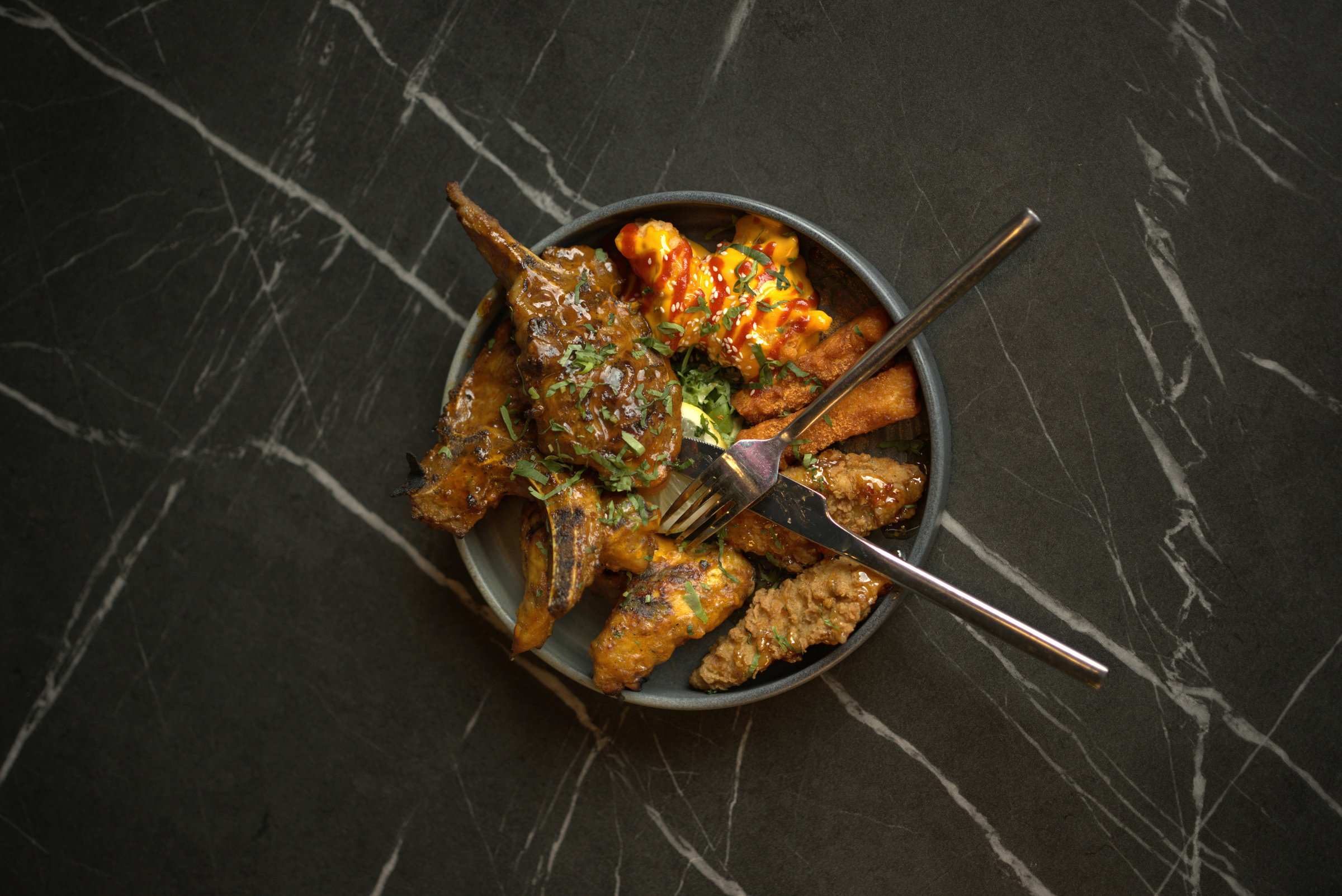 An overhead photograph of glazed chicken wings, garnish and cutlery in a dark bowl.