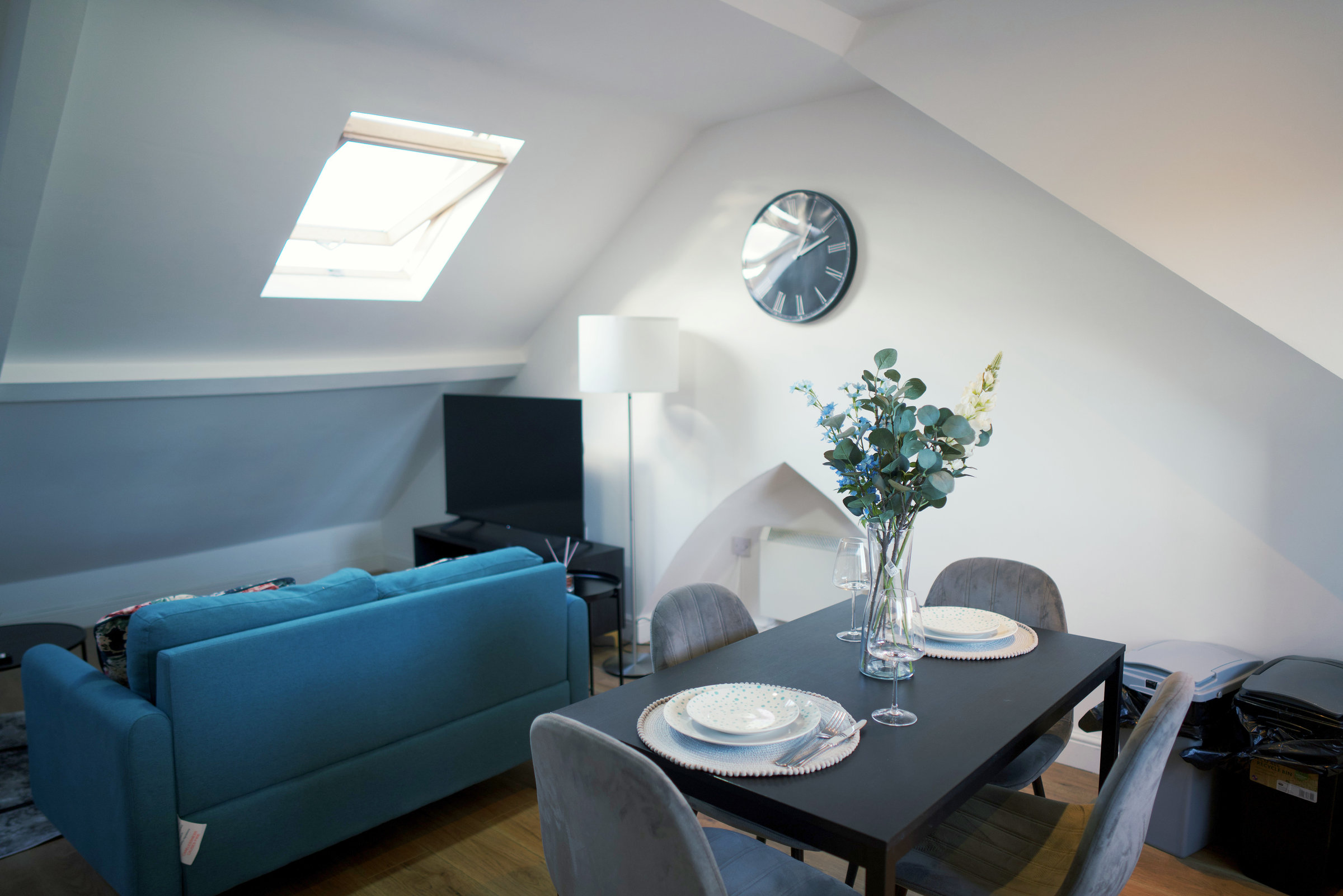 A teal sofa and dining table beneath a skylight in a furnished loft apartment.
