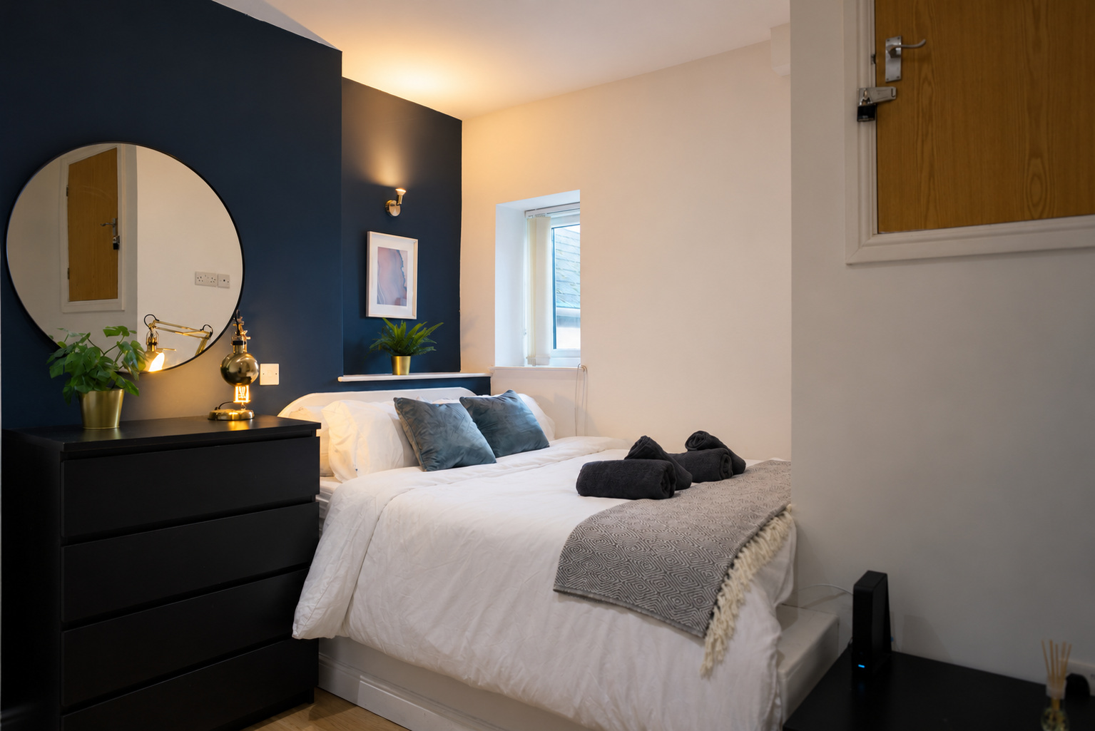 A white bed with blue cushions beside a dark dresser, round mirror and warm brass lamps against a navy wall.