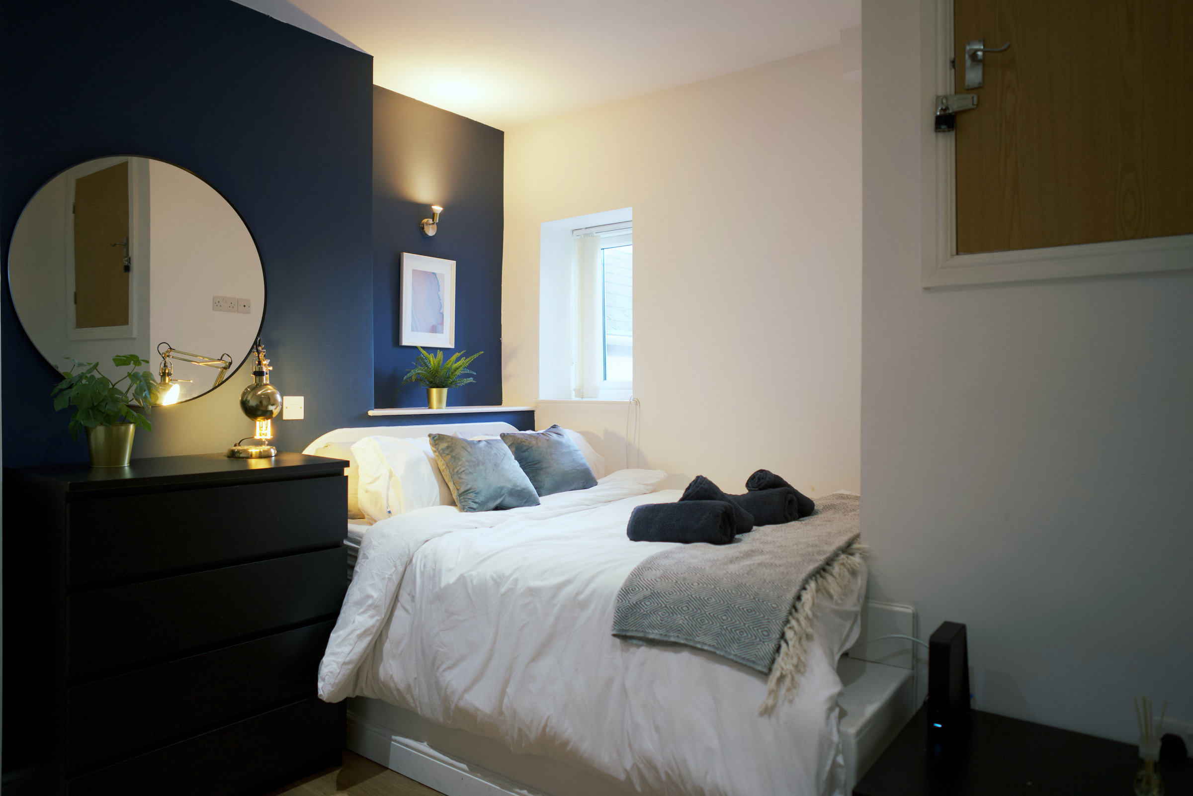 A guest bedroom with a navy feature wall, round mirror and softly lit white bed linen.
