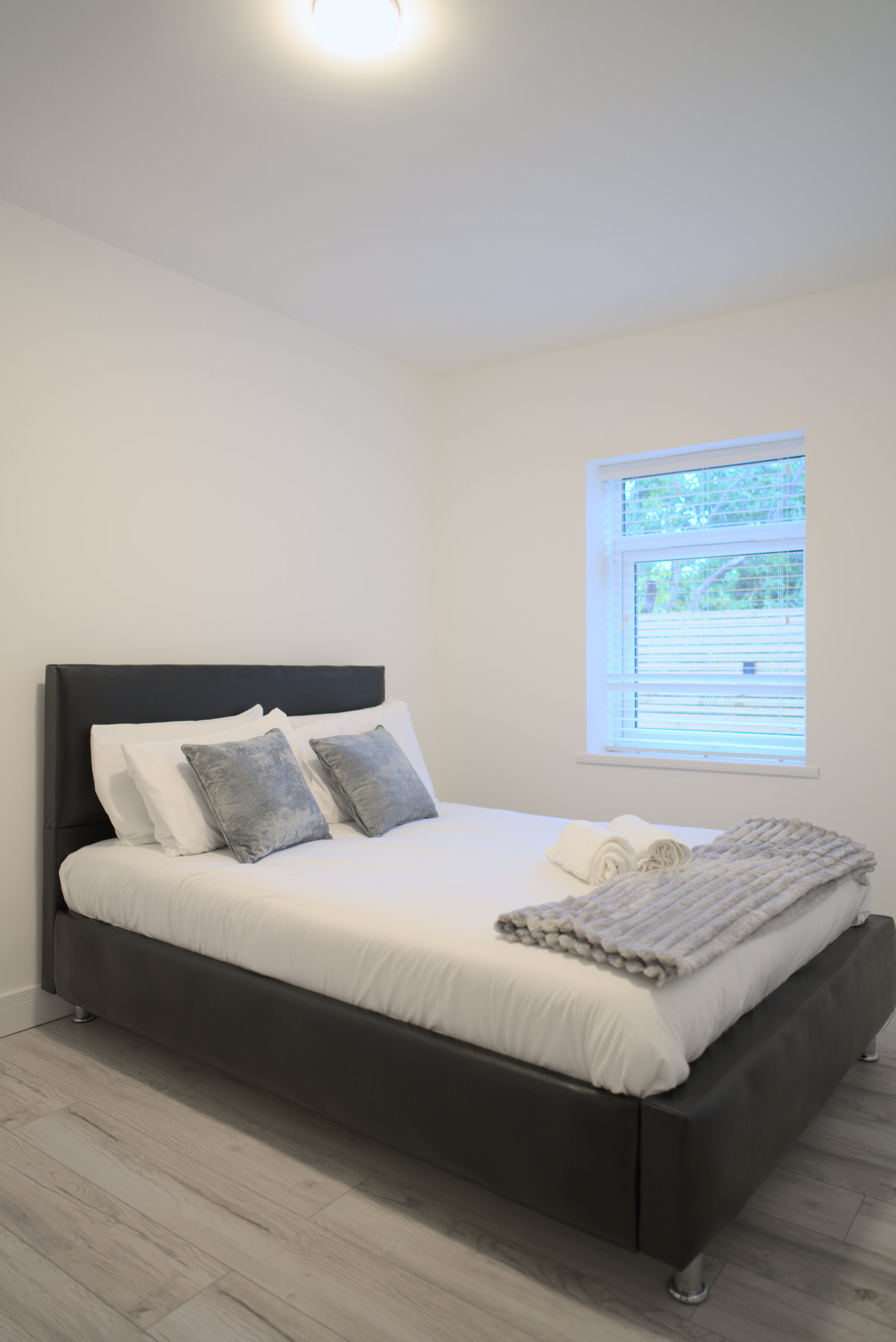 Double bed with grey cushions, folded towels and a window with white blinds.