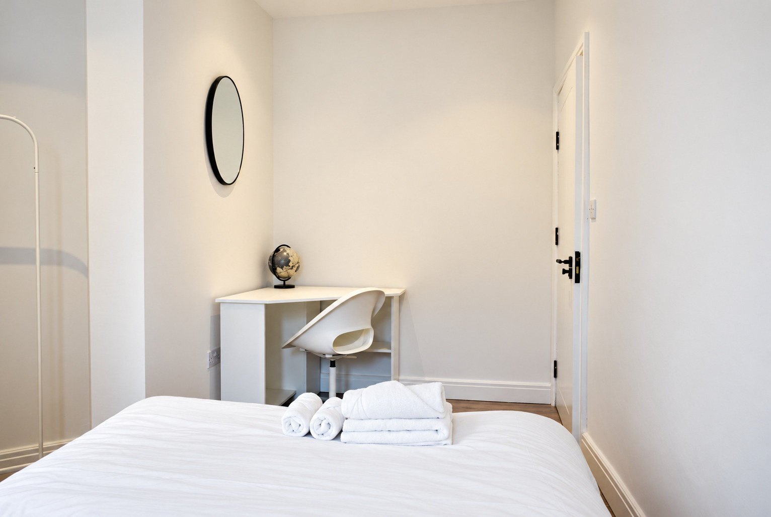 Work desk and round mirror beside a white double bed and rolled towels.