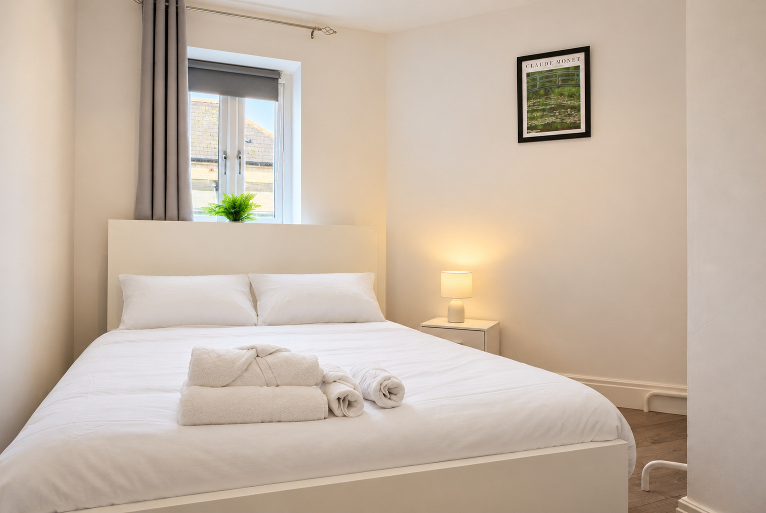 White double bed, rolled towels and a warm bedside lamp beside a bedroom window.