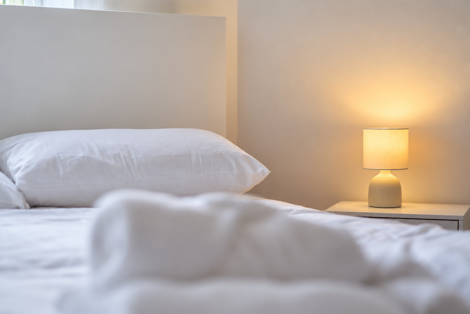 Soft white bed linen in the foreground and a warm bedside lamp beyond.