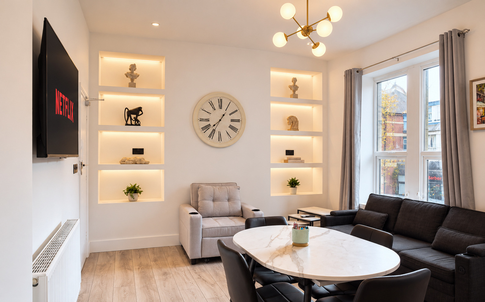 Black sofa and round dining table beside illuminated wall niches and a tall living-room window.