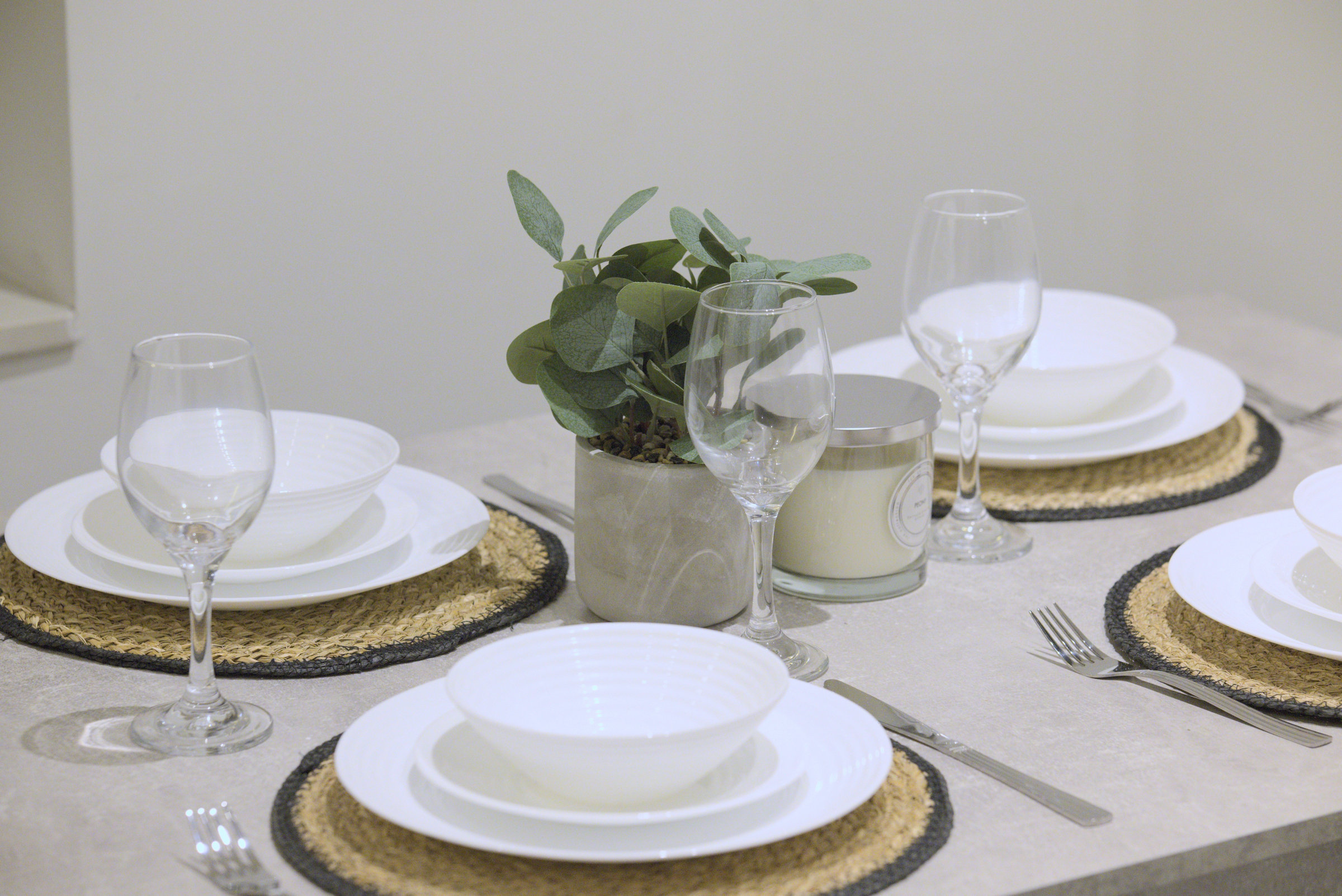 White tableware, clear glasses and woven placemats around a green plant on a dining table.