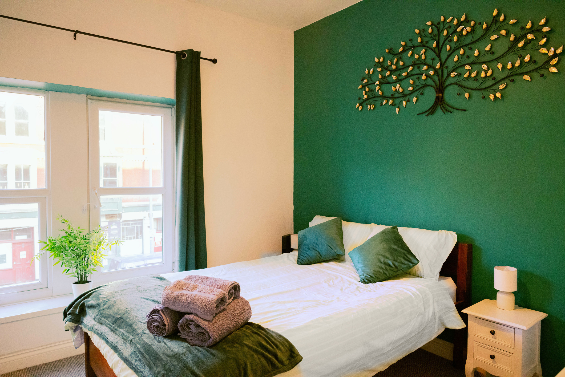White double bed and rolled towels against a green feature wall, with daylight from the adjoining window.