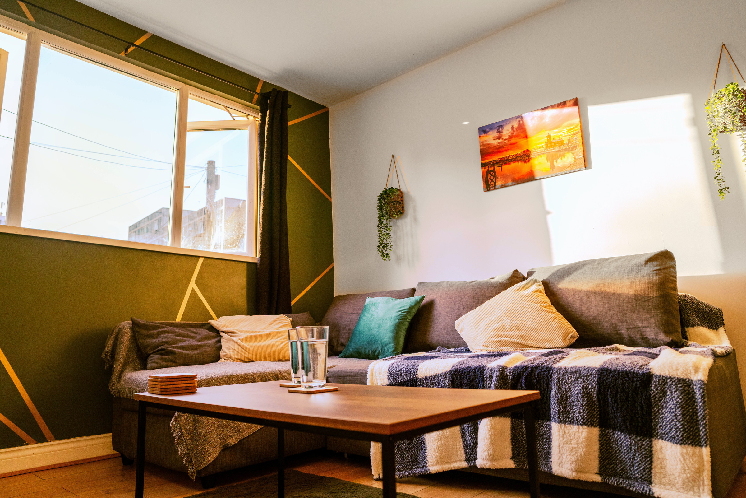 Sunlight across a grey sofa, low wood tables and a green feature wall beside the apartment window.