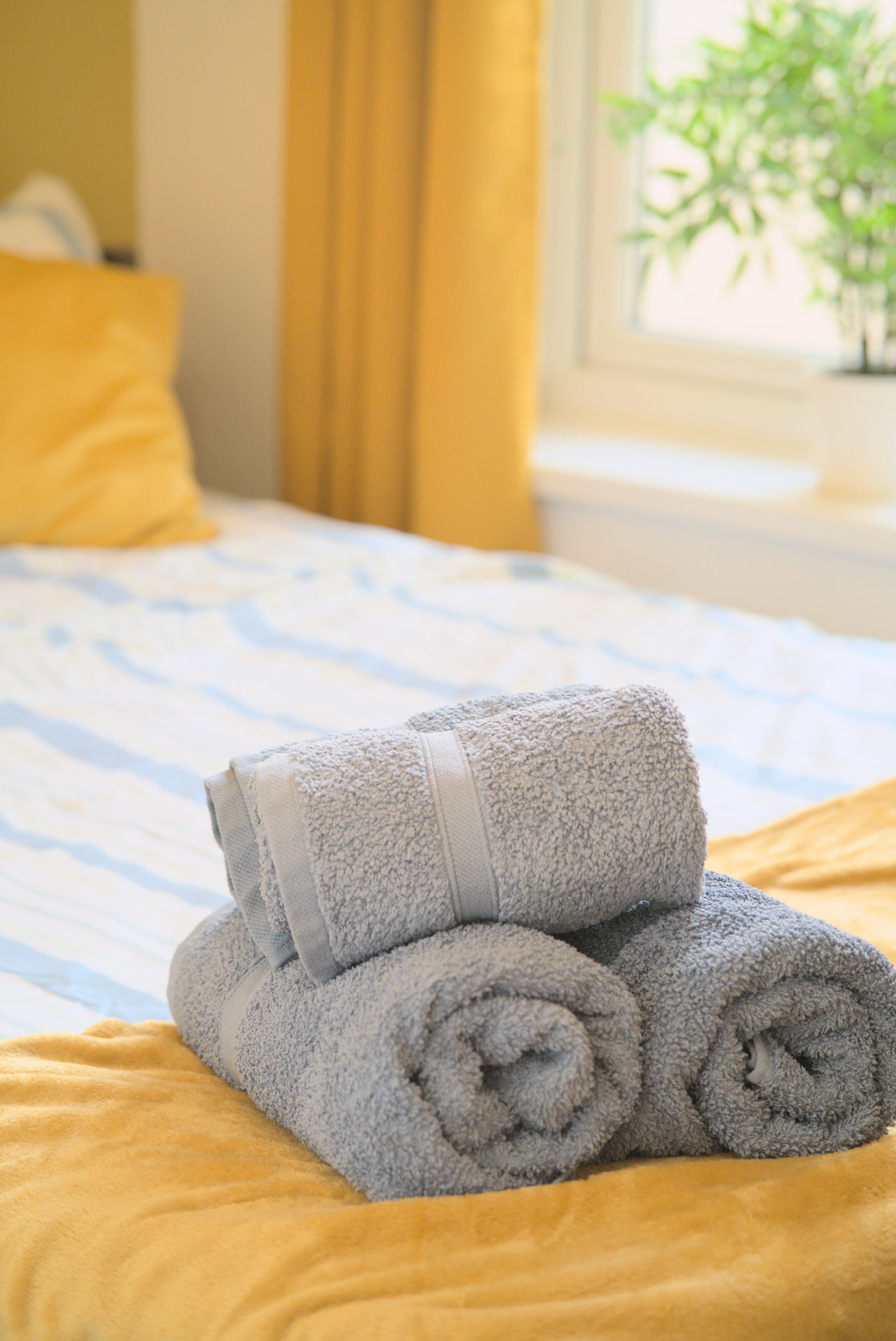 Rolled grey towels on yellow bed linen with sunlight and a plant by the window.