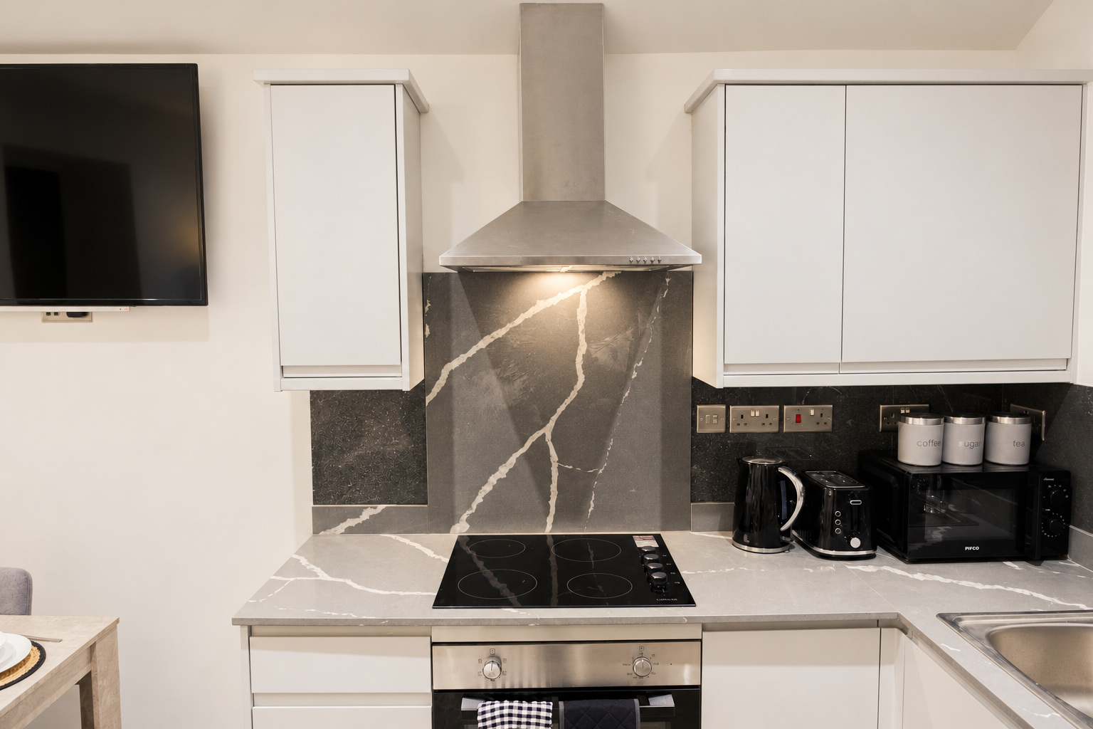 A white apartment kitchen with a black worktop, stainless oven and soft warm light.